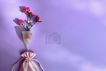 Téléchargez les photos : Coupe menstruelle avec fleurs rouges et sac en soie rose, couché à plat sur fond violet, prise de vue aérienne. Gynécologie et hygiène, zéro déchet alternatives durables. - en image libre de droit