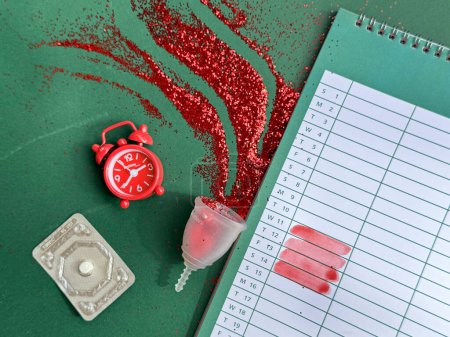 Foto de Taza menstrual con brillo rojo, despertador, píldora anticonceptiva y calendario con días de menstruación marcados en color rojo, concepto de salud femenina vista aérea sobre fondo de papel verde. - Imagen libre de derechos
