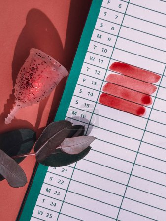 Foto de Copa menstrual con brillo rojo y calendario con días de menstruación marcados en color rojo, imagen concepto de salud femenina. - Imagen libre de derechos
