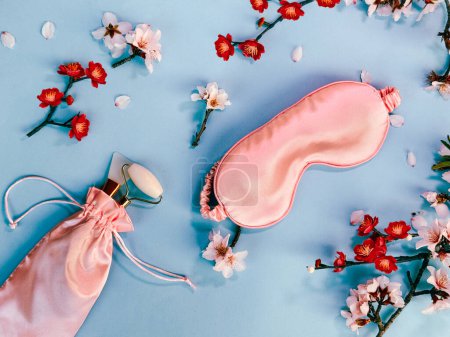 Photo for Quality of sleep, and self care routine. Sleeping mask, massage stone roller in in pink silk bag with spring flowers on blue paper background, overhead background shot. - Royalty Free Image