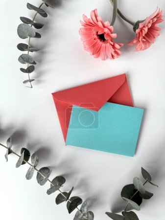 Téléchargez les photos : Une enveloppe bleue et rouge se trouve sur le dessus d'une table, créant une image visuellement frappante avec des fleurs de gerbera rouges. Copier l'espace sur la carte de vœux. - en image libre de droit