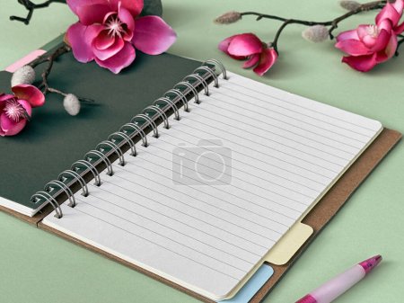 Ein Foto von einem Notizbuch mit einem Stift daneben, zusammen mit einem lebendigen Arrangement von Blumen, die alle auf einem Tisch sitzen. Rosa Magnolienblüten.