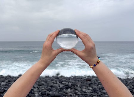 Téléchargez les photos : Mains tenant une boule de cristal sur le bord de l'océan, méditation, voyage spirituel, connexion avec la nature. - en image libre de droit