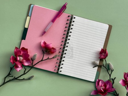 A pink notebook, with its pages neatly bound, is placed at the center of the frame with a pen lying on top of it. Pink magnolia flowers.