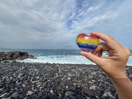 Foto de Mano femenina sosteniendo el corazón de piedra de color arco iris en la orilla del océano, meditación, viaje espiritual, conexión con la naturaleza - Imagen libre de derechos