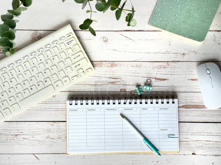 Téléchargez les photos : Cette photo montre un bureau avec un planificateur hebdomadaire vierge, clavier et souris à l'avant-plan, accompagné d'une plante verte. - en image libre de droit