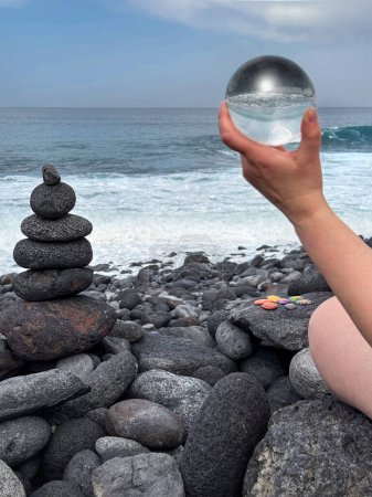 Téléchargez les photos : Main tenant boule de cristal, pyramide zen et pierres de guérison chakra sur le rivage de l'océan volcanique. - en image libre de droit