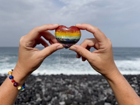 Foto de Manos sosteniendo el corazón de piedra de color arco iris en la orilla del océano, meditación, viaje espiritual, conexión con la naturaleza - Imagen libre de derechos