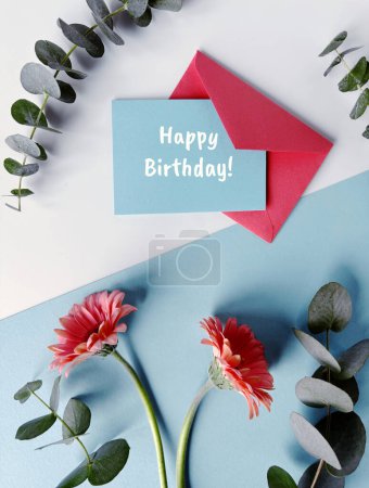 Foto de Una tarjeta de cumpleaños en un sobre azul sobre un fondo de papel de color con vibrantes flores de gerberas rojas. - Imagen libre de derechos