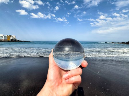Photo for Hand holding glass crystal ball on volcanic sandy beach, ocean shore with waves on a bright day. Meditation, spiritual journey, connection with Nature - Royalty Free Image