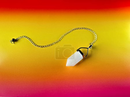 Photo for White crystal pendant, pendulum on a chain, healing amulet, spiritual energy detector on vibrant orange and red paper background. - Royalty Free Image