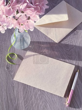 Téléchargez les photos : Un vase rempli de fleurs roses se trouve à côté d'un morceau de papier sur une table. Copier l'espace sur la carte de vœux vierge. - en image libre de droit