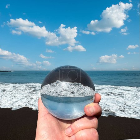 Photo for Close-up on a hand holding glass crystal ball on volcanic seaside, ocean shore with waves on a bright day. - Royalty Free Image
