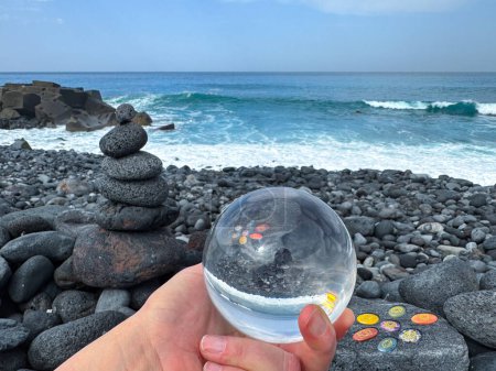 Photo for Hand holding crystal ball, zen pyramid and chakra healing stones on volcanic ocean shore. - Royalty Free Image