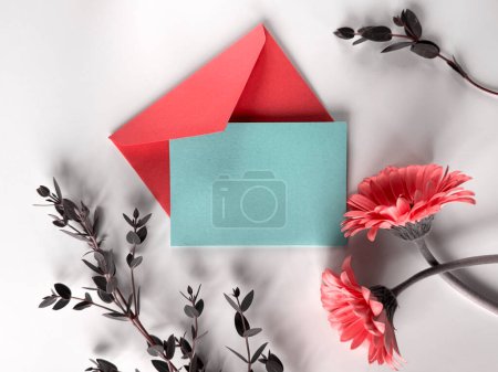 Rote Gerbera-Blüten und ein blauer Umschlag auf einer weißen Oberfläche bilden eine kontrastreiche und lebendige Komposition. Kopierraum auf der Grußkarte.