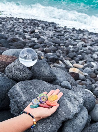 Photo for Healing crystal and hand holding rainbow gemstones, chakra stones, on grey volcanic ocean shore. - Royalty Free Image