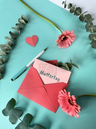 Téléchargez les photos : Une enveloppe rouge avec une note Muttertag, ou fête des mères en langue allemande, attachée à celle-ci, couchée sur une surface verte menthe. - en image libre de droit