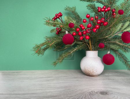 Foto de Arreglo de invierno con ramitas de abeto, decoraciones navideñas y bayas rojas en la mesa de madera junto a la pared verde. - Imagen libre de derechos