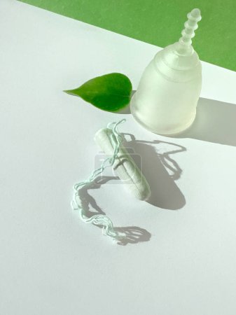 A reusable silicone menstrual cup and standard tampon on white and green paper background, overhead view