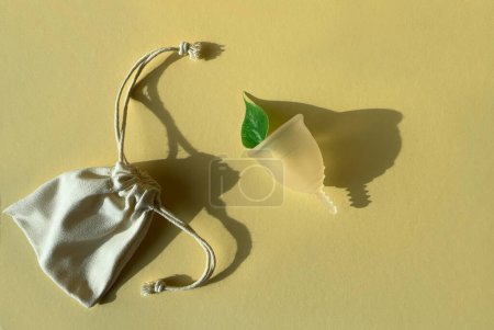 Téléchargez les photos : Coupe menstruelle en silicone réutilisable avec feuille verte sur fond de papier de couleur jaune doré, présentant un produit menstruel unique et respectueux de l'environnement. Couché plat, vue aérienne. - en image libre de droit