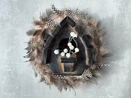 Foto de Corona de plumas de codorniz y casa de pájaros de madera oscura con flores blancas, composición de círculo, decoración de primavera, vista aérea sobre fondo de piedra de hormigón gris. - Imagen libre de derechos