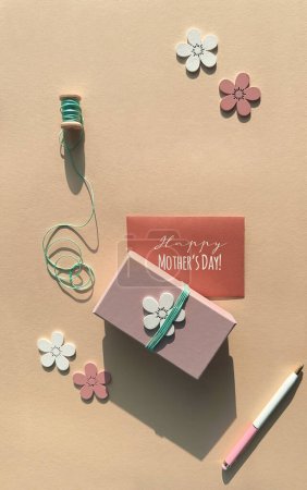 Foto de Tarjeta de felicitación del día de la madre, caja de regalo rosa adornada con flores de madera, carrete de cuerda de cian, pluma sobre papel naranja. - Imagen libre de derechos