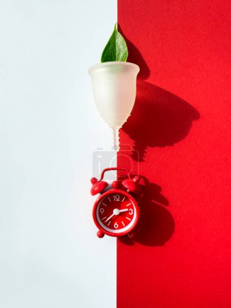 A reusable silicone menstrual cup and alarm clock on a red and white paper, overhead view