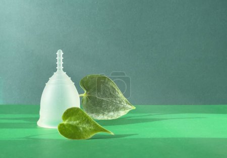 Eine wiederverwendbare Menstruationstasse aus Silikon mit grünem Blatt auf grünem Papierhintergrund, die ein einzigartiges und umweltfreundliches Menstruationsprodukt präsentiert.