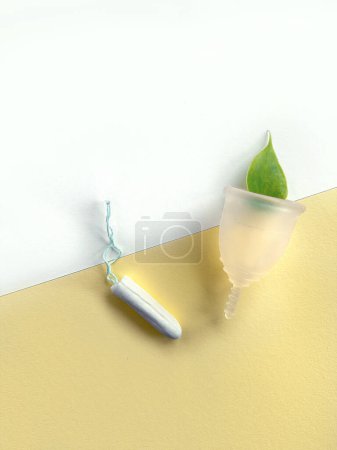 A reusable silicone menstrual cup and standard tampon on white and yellow paper background, overhead view