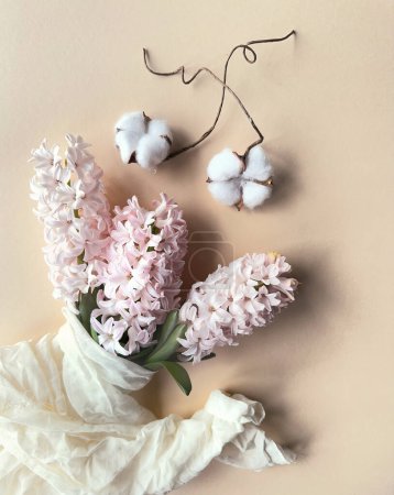 Photo for Spring composition with pink hyacinth wrapped in linen towel and white cotton flowers, overhead view on cream, beige paper background. - Royalty Free Image