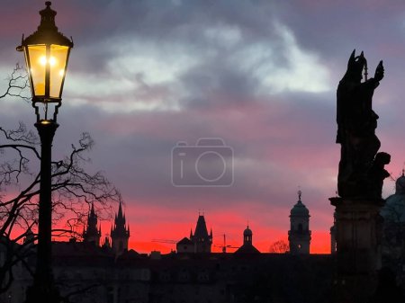 Photo for Dawn in Prague: Charles Bridge with pink-purple clouds, statue silhouette, and glowing street light. - Royalty Free Image