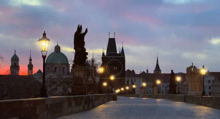Morgendämmerung in Prag: Karlsbrücke mit rosa-violetten Wolken, Statuensilhouetten und leuchtenden Straßenlaternen.