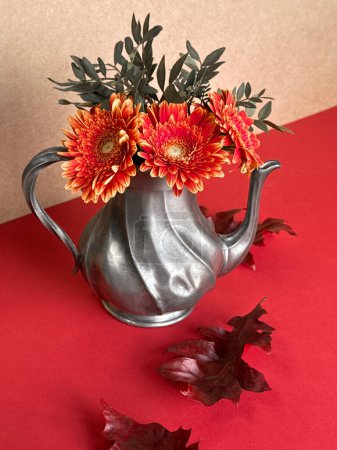 Vintage-Metall-Teekanne mit leuchtend orangen Gerbera-Blüten und Herbstblättern auf rotem Hintergrund.