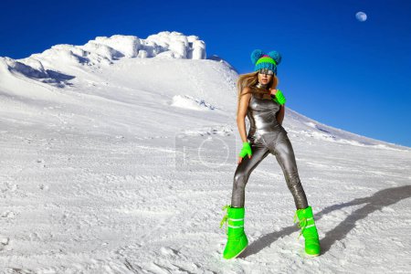 Sexy Frau in silbernem Kosmos-Outfit und Winterstiefeln posiert in den Bergen. Weibliches Model im silbernen Glamour-Jumpsuit in Winterkulisse.