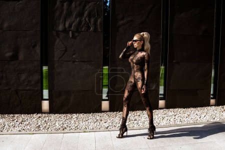 Foto de High fashion look. Glamour stylish model in black latex clothing and and high heels on is walking in the city. Sexy stylish blonde woman in bodystocking lingerie is posing outdoor. Sexy Lingerie model. - Imagen libre de derechos