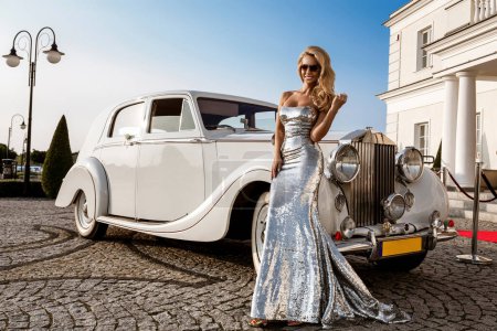 Una hermosa mujer estrella en un vestido de noche largo y lujoso plateado se encuentra cerca de un coche vintage y posa sensualmente.