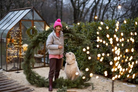Foto de Mujer joven juega con su adorable perro, divirtiéndose juntos mientras están de pie con una gran corona de Navidad en el patio trasero. Concepto de amor y comodidad durante las vacaciones de invierno - Imagen libre de derechos