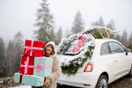 Eine Frau hält einen Haufen Geschenkboxen in der Hand, während sie neben dem mit Weihnachtskranz und Baum geschmückten Auto auf dem Dach steht, das bei verschneitem Wetter mit dem Auto in den Bergen unterwegs ist. Konzept der Winterferien
