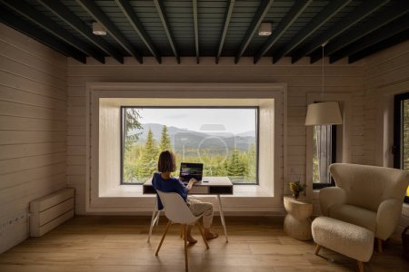 Photo for Woman works on laptop while sitting by the table in front of panoramic window with great view on mountains. Wide interior view. Remote work and escaping to nature concept - Royalty Free Image