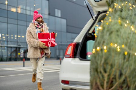 Photo pour Jeune femme gaie en vêtements d'hiver se tient avec une boîte-cadeau près du centre commercial et l'arbre de Noël en face. Concept de shopping pour les vacances d'hiver - image libre de droit