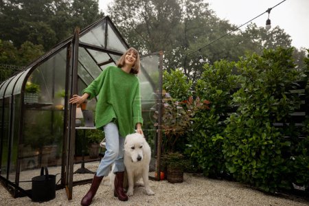 Téléchargez les photos : Jeune femme avec son chien blanc près de la serre à l'arrière-cour, passer du temps libre ensemble à l'extérieur - en image libre de droit