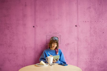 Young stylish woman works on a digital tablet while sitting on pink concrete wall background at modern coffee shop. Concept of remote creative work online