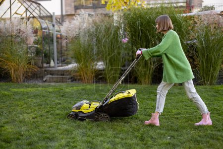 Foto de Woman cuts grass with electric lawn mower on backyard of her country house. Garden care and modern garden tools concept - Imagen libre de derechos