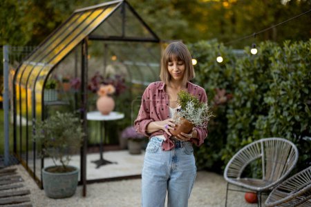Téléchargez les photos : Jeune femme jardinier porte pot de fleurs avec des herbes sur la cour arrière avec une belle serre pour les plantes sur fond - en image libre de droit