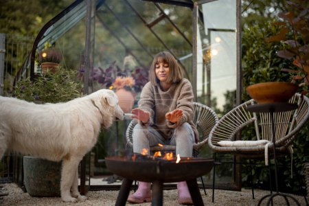 Téléchargez les photos : Jeune femme se réchauffe les mains, passer du temps en soirée avec son chien au coin du feu dans un jardin confortable et magnifique - en image libre de droit