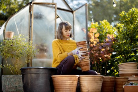 Foto de Woman plants flowers in clay jugs, sitting in front of glass orangery at garden in sunny morning. Gardening and hobby concept - Imagen libre de derechos