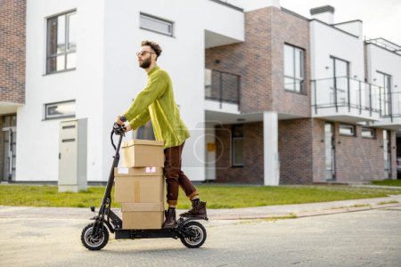 Téléchargez les photos : Homme conduit scooter électrique, boîtes de carton de livraison sur la rue dans un quartier résidentiel. Concept de durabilité, mode de vie moderne et respectueux de l'environnement - en image libre de droit