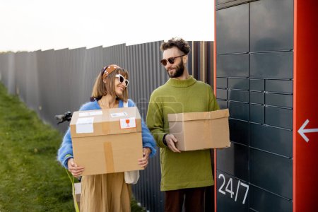 Téléchargez les photos : Joyeux jeune homme et femme portent des colis en carton reçus ensemble dans la machine automatique de courrier à l'extérieur. Concept de technologies de livraison modernes - en image libre de droit