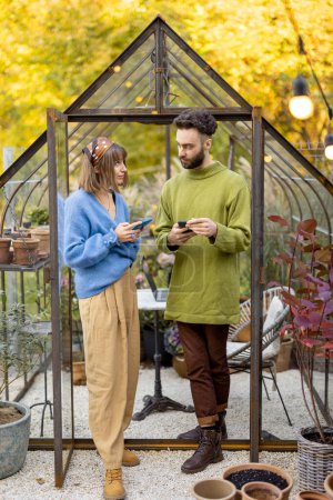 Photo for Young stylish man and woman talk together while standing with phones in front of tiny glass orangery in garden. Two hipsters hang out outdoors - Royalty Free Image