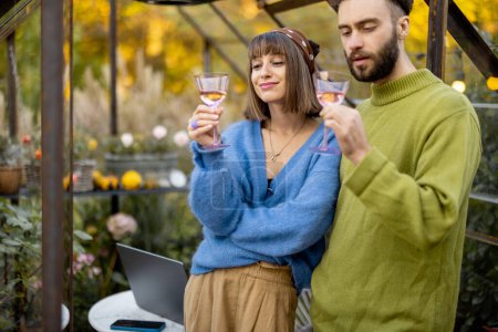 Photo for Young stylish couple talk and drink wine, spending leisure time together near glass house in garden. Man and woman have romantic eve time with alcohol drink at backyard - Royalty Free Image